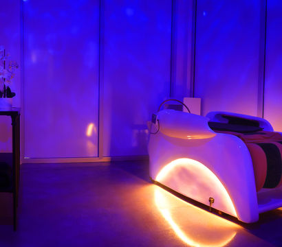 Futuristic white relaxation pod in a dim spa room with blue-purple ambient lighting, warm orange underglow and an orchid on a side table