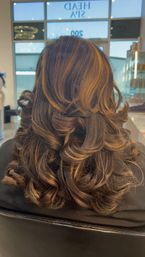 Back view of shoulder-length dark brown hair with warm caramel highlights, styled into voluminous layered curls and flipped ends in a salon.