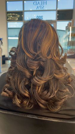 Back view of shoulder-length dark brown hair with warm caramel highlights, styled into voluminous layered curls and flipped ends in a salon.