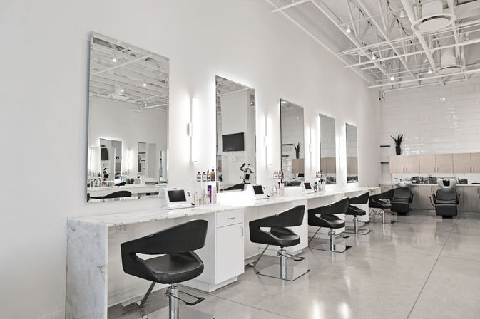 Bright modern hair salon interior with a row of illuminated mirrors and marble-topped styling stations, black swivel chairs, shampoo sinks, high exposed ceiling, and polished concrete floor.