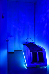 Glowing blue LED relaxation pod in a dim spa treatment room, sleek massage/hydrotherapy bed with curtain backdrop