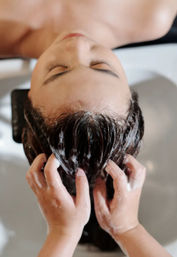 Top-down view of a relaxed salon client reclining with eyes closed as a stylist massages soapy shampoo into wet hair over a salon wash basin.