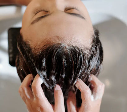 Top-down view of a relaxed salon client reclining with eyes closed as a stylist massages soapy shampoo into wet hair over a salon wash basin.