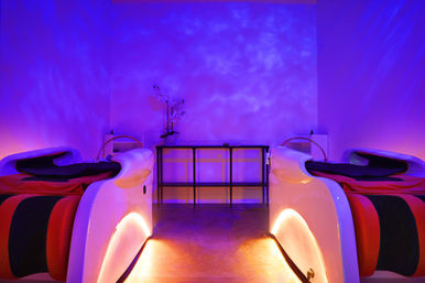 Two futuristic white spa pods with red-and-black cushions in a dim purple-lit relaxation room, warm underlighting and an orchid on a central console — calming wellness spa ambience.