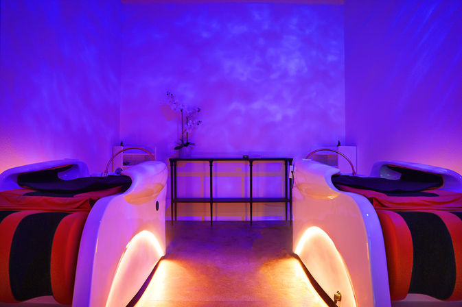 Two futuristic white spa pods with red-and-black cushions in a dim purple-lit relaxation room, warm underlighting and an orchid on a central console — calming wellness spa ambience.