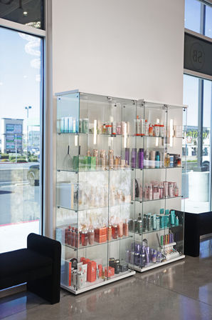 Bright salon interior with glass display case of colorful hair and skincare bottles and tubes on retail shelves by large street-facing windows and black seating