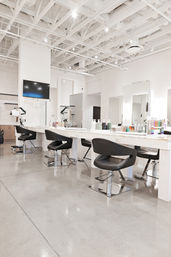Bright modern hair salon interior with white exposed-beam ceiling, polished concrete floor, long white styling stations with large mirrors, black swivel chairs and product displays