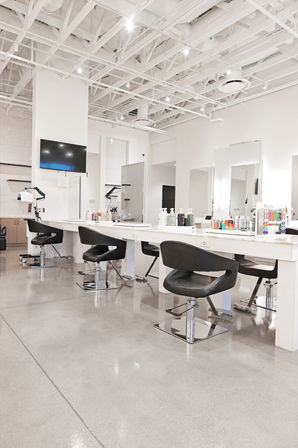Bright modern hair salon interior with white exposed-beam ceiling, polished concrete floor, long white styling stations with large mirrors, black swivel chairs and product displays