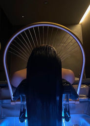 Person reclining at a salon spa hair-wash station, long wet hair cascading under a semicircular rainfall shower into a blue-lit basin