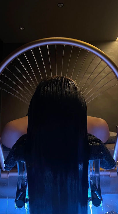 Person reclining at a salon spa hair-wash station, long wet hair cascading under a semicircular rainfall shower into a blue-lit basin