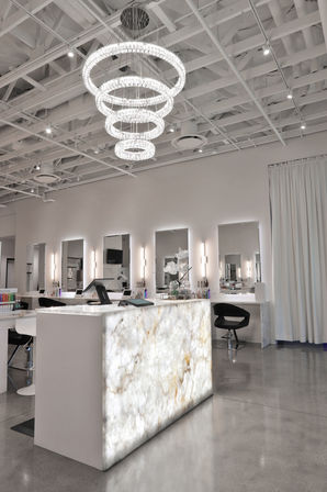 Modern white salon interior with illuminated marble reception desk, tiered crystal chandelier, mirrored styling stations and polished concrete floor.