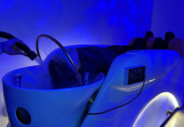 Relaxing scalp steam treatment in a white hydrotherapy spa pod with blue LED mood lighting, digital control panel, neck support and a handheld steaming nozzle