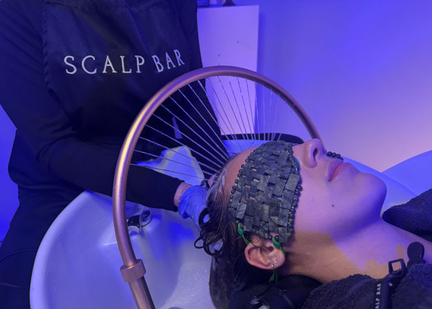 Client reclining at a salon sink receiving a scalp treatment under a copper arched ring with fine water jets, wearing a woven stone eye mask and bathed in blue LED light.