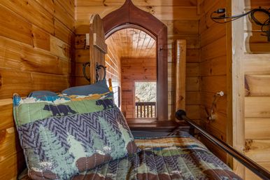 Cozy rustic wood-paneled cabin bunk with forest-and-bear quilt and pillow, arched wooden doorway opening to a small balcony overlooking trees.