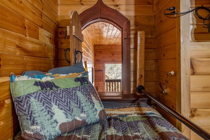 Cozy rustic wood-paneled cabin bunk with forest-and-bear quilt and pillow, arched wooden doorway opening to a small balcony overlooking trees.