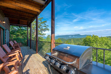Wooden mountain cabin deck with rocking chairs and a gas grill overlooking lush forested hills and a blue-sky mountain panorama