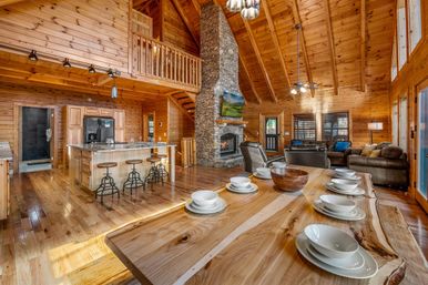 Sunlit rustic mountain cabin interior with vaulted wood ceilings, stone fireplace and loft; open-plan kitchen with island and metal bar stools, and a live-edge dining table set with white dishes.
