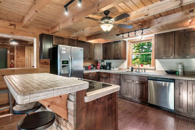 Rustic wood cabin kitchen with exposed beam ceiling, tile-topped island and bar stools, stainless steel fridge and dishwasher, dark wood cabinets, ceiling fan and window view of trees.