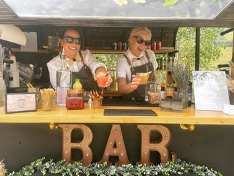 Two smiling bartenders in aprons and sunglasses serving colorful cocktails from a rustic outdoor mobile bar with a large "BAR" sign and greenery