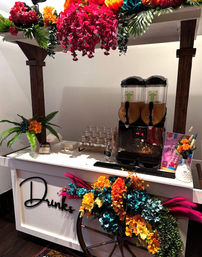 Indoor event drink cart with dual beverage dispensers and stacked glasses, white wooden 'Drinks' sign, rustic wagon wheel and vibrant teal, orange, pink and yellow floral arrangements for a colorful beverage station.