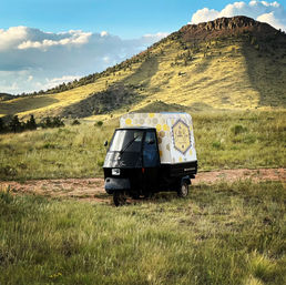 Tiny black three-wheeled delivery vehicle with a yellow honeycomb cargo box parked in a grassy mountain meadow beneath a sunlit rocky butte and blue sky.