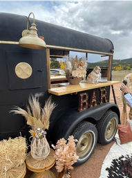 Vintage black trailer converted into a mobile bar with a wooden serving counter and marquee 'BAR' sign, a small tan chihuahua perched on the counter, dried floral arrangements, pampas grass, woven baskets and hay bales set in an outdoor autumn meadow with yellow trees and cloudy sky.