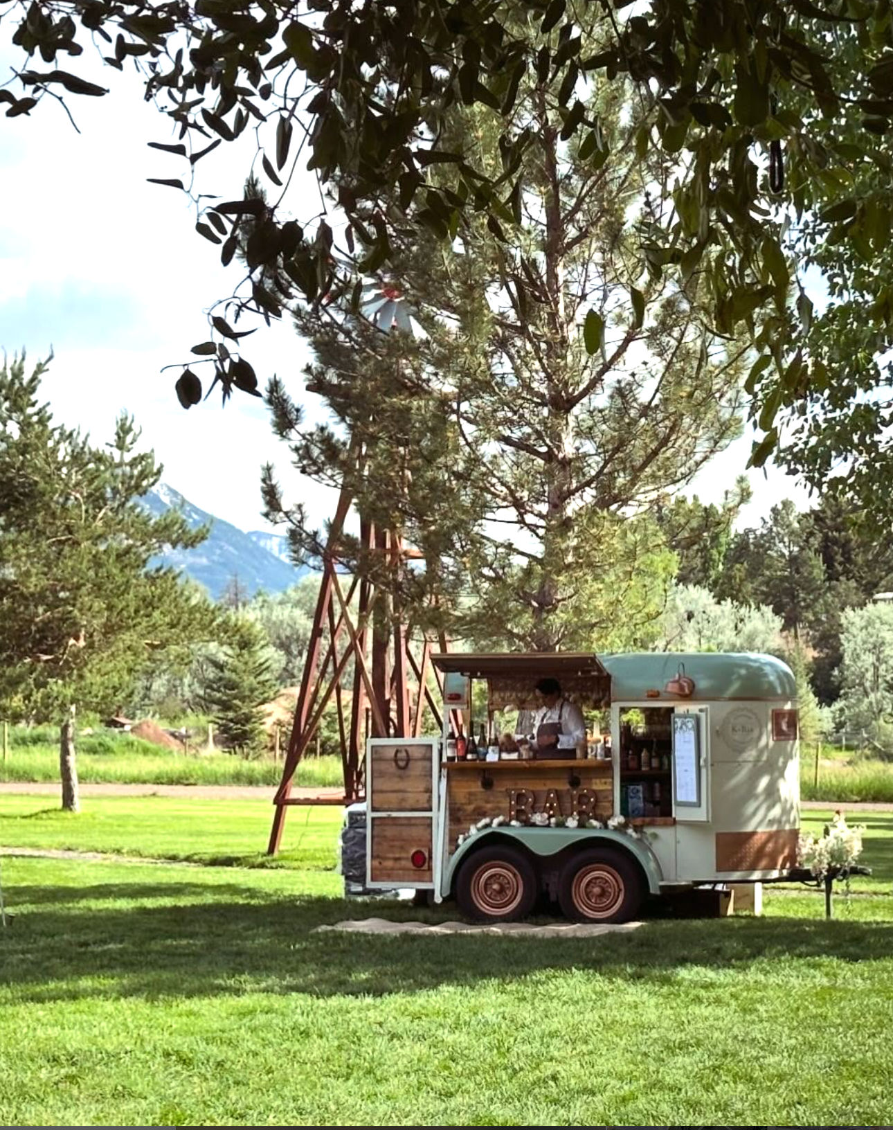 Charming vintage mobile bar trailer on a sunlit grassy lawn beneath pine trees, bartender serving from a wooden counter with a rustic windmill and distant mountains in the background.