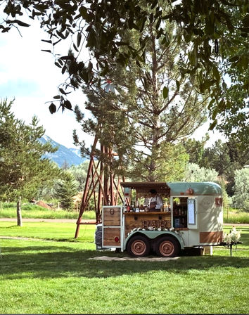 Charming vintage mobile bar trailer on a sunlit grassy lawn beneath pine trees, bartender serving from a wooden counter with a rustic windmill and distant mountains in the background.
