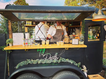 Outdoor mobile bar trailer with wooden counter, two servers in white shirts and leather aprons preparing cocktails; jars of dried citrus, canned mixers and greenery garland decorate the setup for an outdoor event.