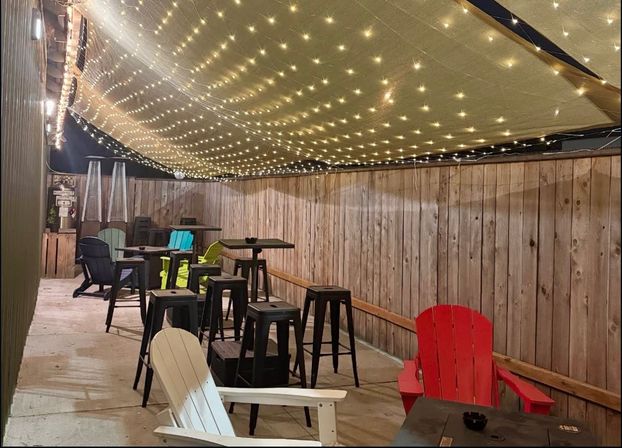 Cozy fenced outdoor patio with a canopy of twinkling string lights, colorful Adirondack chairs, high bar stools and tables, and patio heaters for evening seating.