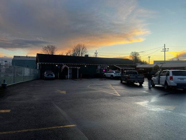 Rain-slick parking lot at dusk in front of a low, single-story building decorated with string lights and an American flag, parked cars and trucks, and a golden-pink sunset sky reflected on the asphalt.