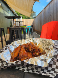 Sticky BBQ chicken wings in a foil-lined basket on black-and-white checkered paper, al fresco patio scene with high-top stools, red chair, sunshade and string lights