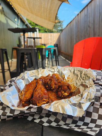 Sticky BBQ chicken wings in a foil-lined basket on black-and-white checkered paper, al fresco patio scene with high-top stools, red chair, sunshade and string lights