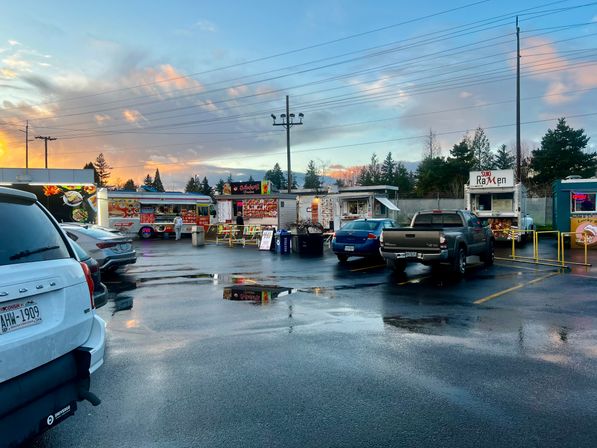 Colorful food trucks in a wet suburban parking lot at sunset, parked cars and puddles reflecting neon signs and pastel clouds — a cozy outdoor food truck scene.