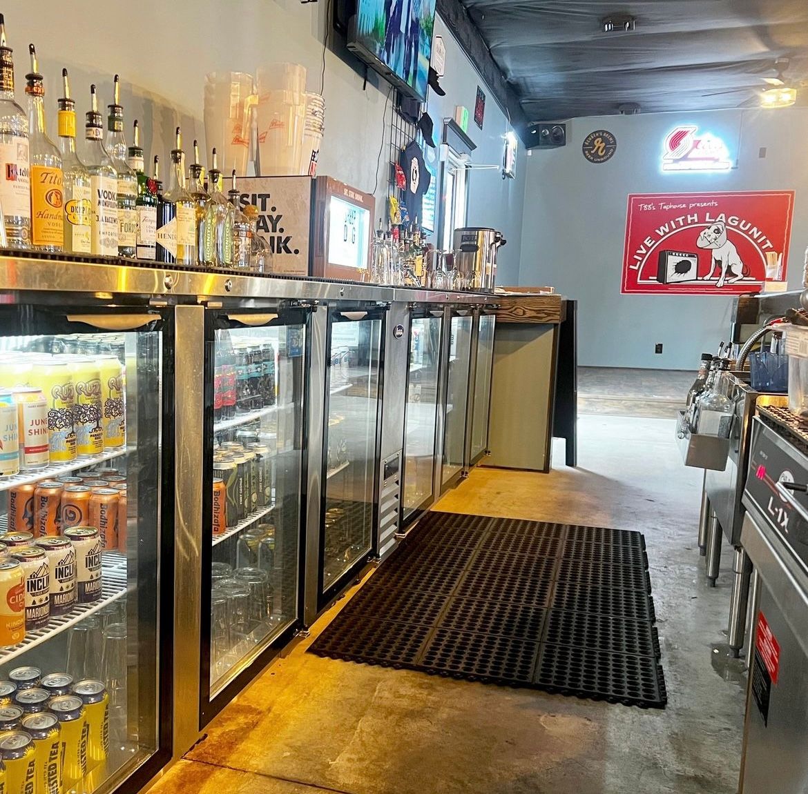 Taproom interior with stainless bar counter, liquor bottles on top and glass-door fridges stocked with craft beer cans, neon beer poster on the back wall and rubber floor mat on concrete floor.