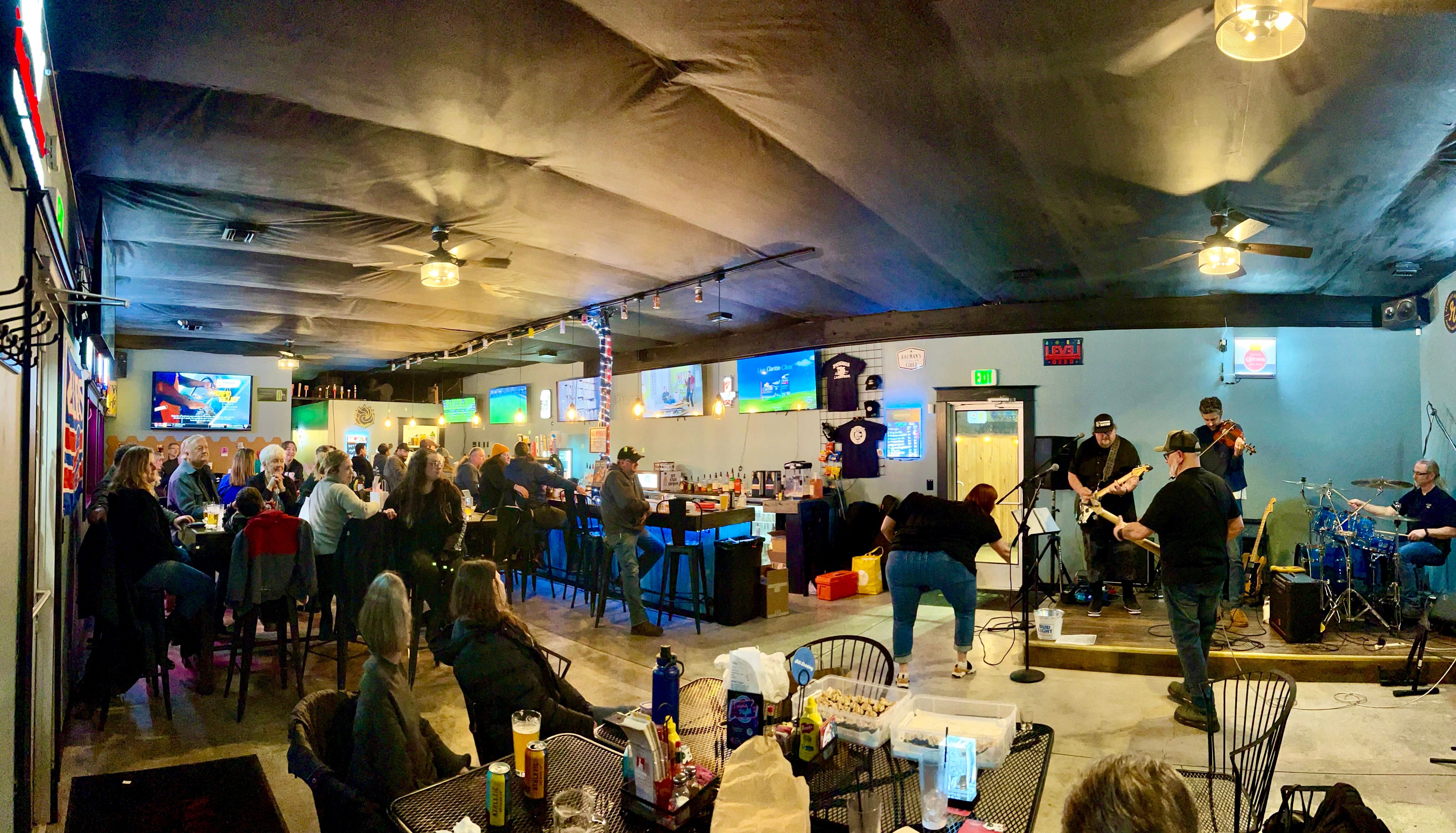 Panoramic interior of a lively neighborhood bar with seated patrons, a central bar and multiple TVs, and a band performing live on a small stage.