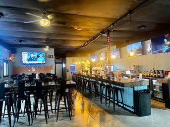 Empty casual sports bar interior with a long wooden bar, rows of black metal barstools and high-top tables, warm pendant lighting, multiple wall-mounted TVs showing sports, concrete floor and industrial ceiling.