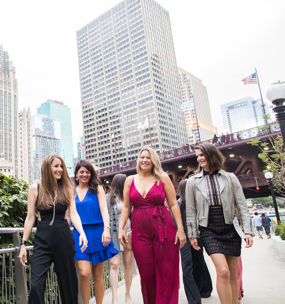 Four smiling women in colorful outfits strolling the Chicago Riverwalk with downtown skyscrapers and a steel bridge rising behind them.