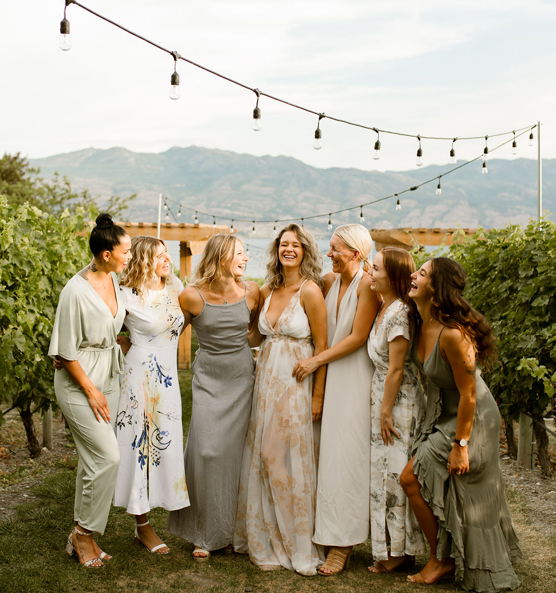 Bridal party of seven laughing together in a vineyard under string lights with mountain views — outdoor wedding vibe