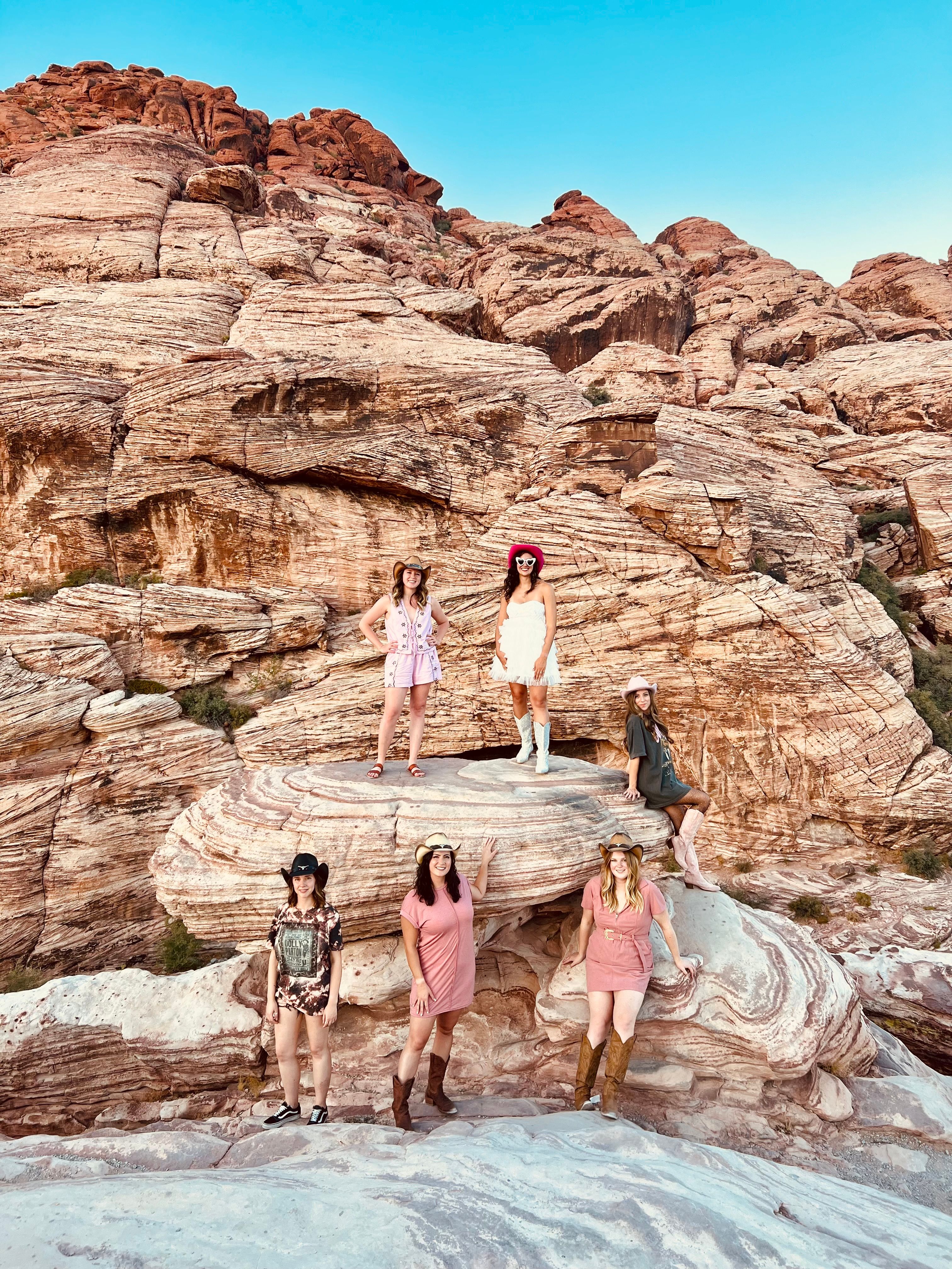 Six women wearing cowboy hats and boots posing on layered red sandstone rock formations in a desert canyon under a clear blue sky.