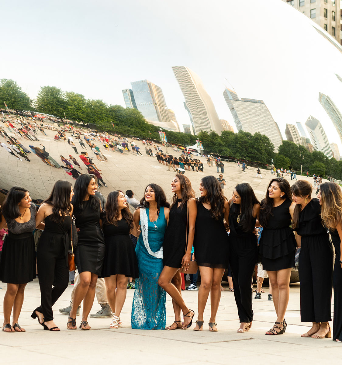 Joyful group of women—mostly in black dresses with one in a blue dress and sash—posing and laughing in front of Chicago’s Cloud Gate (The Bean) with mirrored skyline reflections.