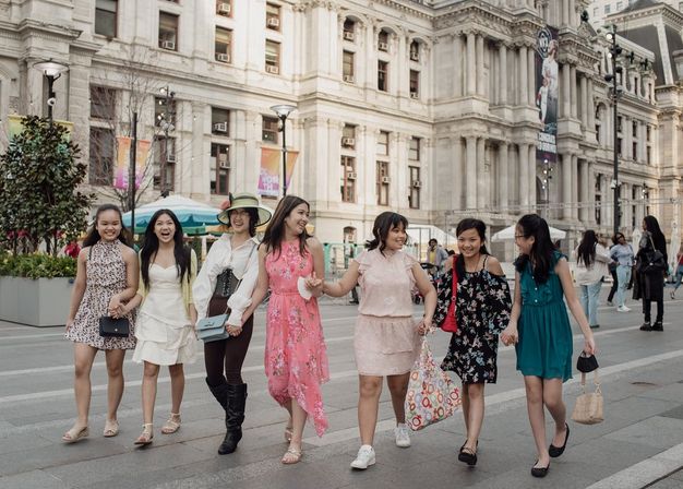 Seven friends in colorful summer dresses walking hand-in-hand through an urban plaza in front of a grand historic stone civic building, enjoying a lively city outing.