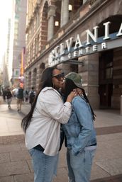 Two people sharing an affectionate hug and kiss on a downtown city sidewalk in front of a historic theater marquee, one in sunglasses and a light coat and the other in a camo cap and denim jacket — warm urban moment.