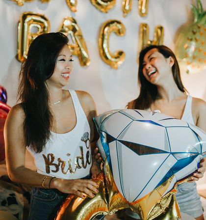 Two friends laughing at a bachelorette party, bride-to-be wearing a 'Bride' tank top and holding a large diamond-shaped foil balloon with gold letter balloons in the background, indoor celebration