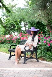 Stylish woman in a purple wide-brim sun hat and light dress sitting on a black park bench, gold heels visible, surrounded by pink roses and lush greenery in a city park garden.
