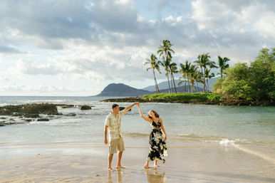 Couple dancing barefoot on a tropical island beach with shallow waves, sandy shore, palm trees, rocky outcrops and a distant mountain under a partly cloudy sky.