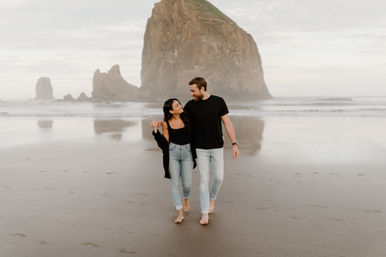 Couple walking barefoot on a reflective sandy beach at Cannon Beach, Oregon, holding hands and smiling with Haystack Rock rising in the misty background
