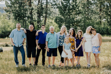 Multigenerational family portrait in a sunny grassy meadow by pine trees — adults, teens and children smiling in casual summer clothing for an outdoor family photo.