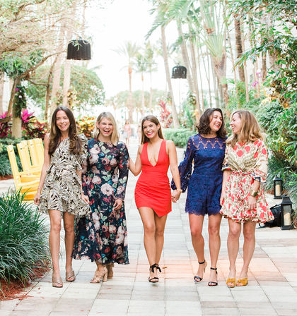 Five women in colorful summer dresses laughing and walking down a palm‑lined tropical resort walkway with lush greenery and bright yellow chairs