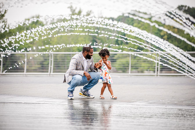 Father and young daughter holding hands and laughing as arched water jets spray over an outdoor urban fountain plaza with wet reflective pavement and trees in the background
